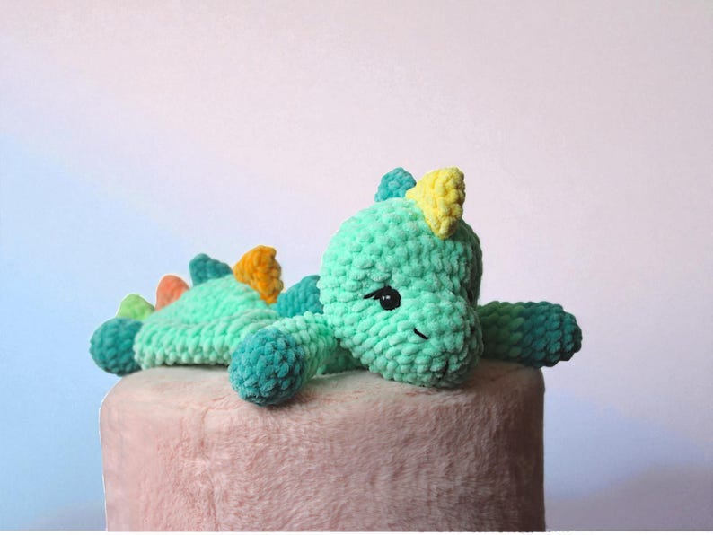 May include: A handmade, plush dinosaur toy in shades of teal, green, yellow, orange, and blue. The dinosaur has a friendly expression with black eyes and is lying on a pink surface. The toy is made of soft, textured yarn.
