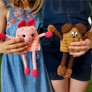May include: Two handmade crochet dolls. One is pink with a bunny-like design, featuring pink ears, arms, and feet. The other is brown, resembling a dog, with brown ears, arms, and feet. Both dolls have black eyes and are held by a person.