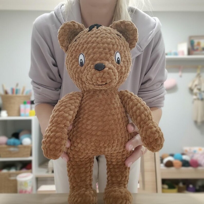 May include: A handmade brown teddy bear with a black nose and black accents on top of its head. The bear has a friendly expression with white and black eyes. The bear is made of a soft, textured material and is approximately 18 inches tall.