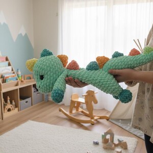 May include: A handmade, plush, green crochet dinosaur toy with colorful accents. The toy has a long body, a cute face, and colorful spikes along its back. The dinosaur is being held in front of a child's room setting.