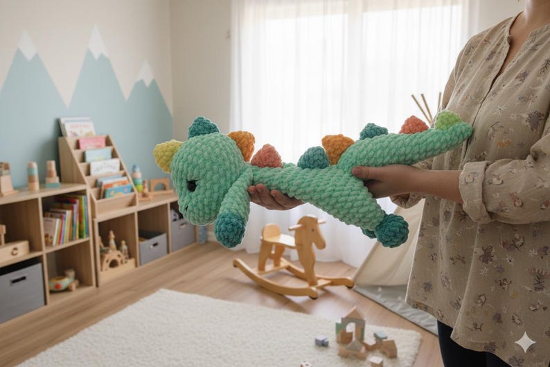 May include: A handmade, plush, green crochet dinosaur toy with colorful accents. The toy has a long body, a cute face, and colorful spikes along its back. The dinosaur is being held in front of a child's room setting.