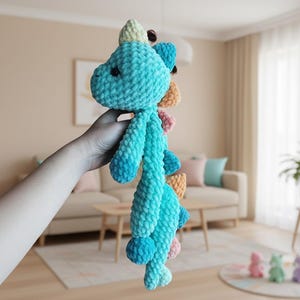 May include: A handmade, plush turquoise dinosaur toy with a light blue belly and colorful spines. The dinosaur is held up in front of a neutral background. The toy is made of soft, textured yarn.