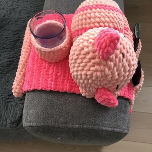 May include: A handmade, pink and light pink crocheted couch arm organizer with a cup holder and a pig-shaped remote control holder. The cup holder is holding a clear glass. The organizer is on a gray couch.