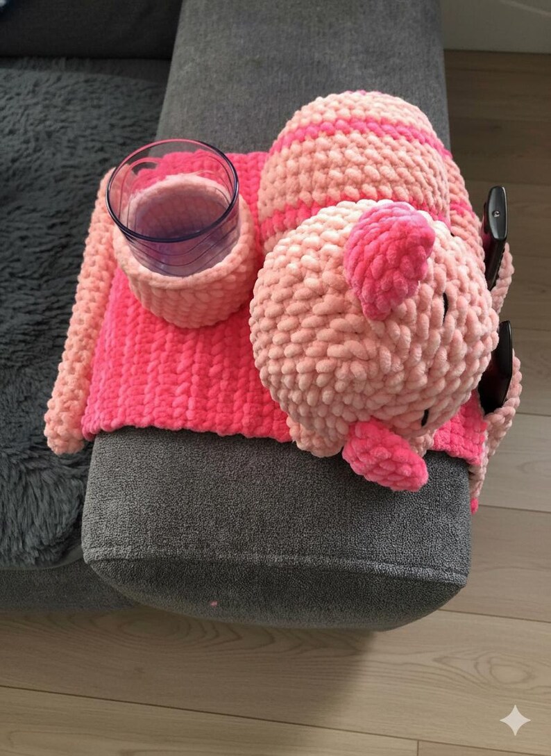 May include: A handmade, pink and light pink crocheted couch arm organizer with a cup holder and a pig-shaped remote control holder. The cup holder is holding a clear glass. The organizer is on a gray couch.