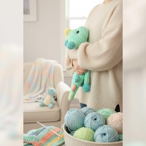 May include: A crocheted dinosaur toy in shades of green, blue, and yellow is held by a person. A basket of yarn in various pastel colors sits in the foreground, along with a crocheted blanket and a small stuffed animal.