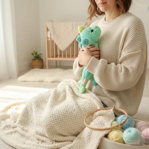 May include: A person holding a crocheted dinosaur toy. The toy is primarily turquoise with yellow and orange accents. A basket with yarn in various pastel colors and a wooden embroidery hoop are in the foreground. A cream-colored blanket and sweater are also visible.