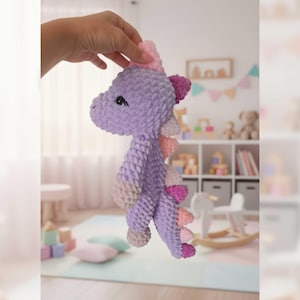 May include: A handmade, lavender crochet dinosaur toy with pink and peach accents. The plush toy has a soft, textured appearance, with small black eyes and a pink horn. The dinosaur's back features a row of pink and peach spikes. A charming, whimsical children's toy.