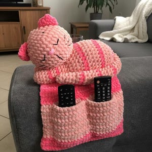 May include: A handmade, pink and fuchsia crochet cat-shaped remote control holder. The cat has a striped body and two pockets for remotes. The cat is lying on a gray couch. The cat's head is at the top, with its eyes closed.