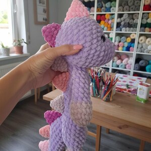 May include: A handmade, plush dinosaur toy in shades of purple, pink, and peach. The soft, textured toy features a spiky back and a friendly expression. This cuddly creature is perfect for children and makes a great gift.