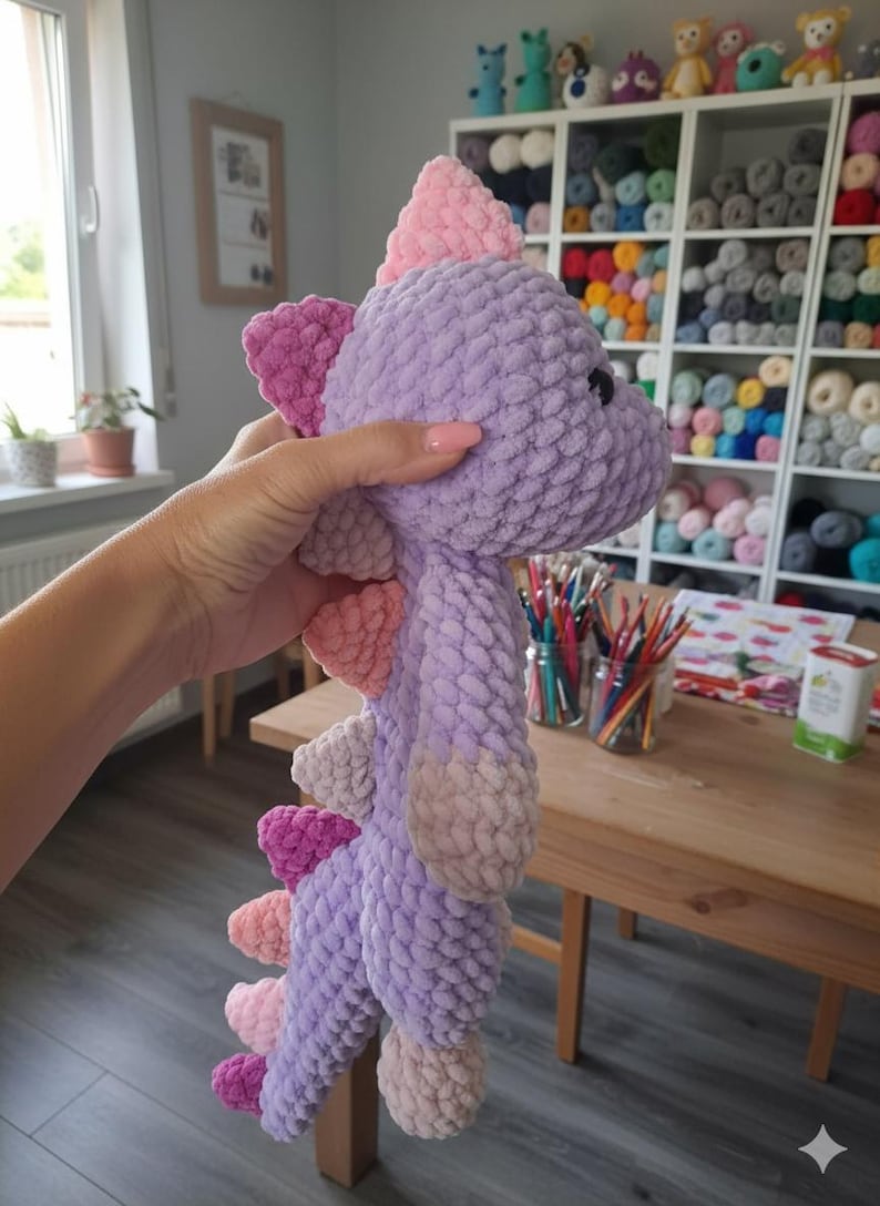 May include: A handmade, plush dinosaur toy in shades of purple, pink, and peach. The soft, textured toy features a spiky back and a friendly expression. This cuddly creature is perfect for children and makes a great gift.