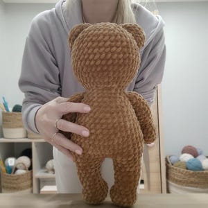 May include: A handmade brown teddy bear made of soft, textured yarn. The bear has a round head, small ears, and a plump body. It is held up by a person. The bear is a classic toy and a cozy gift.