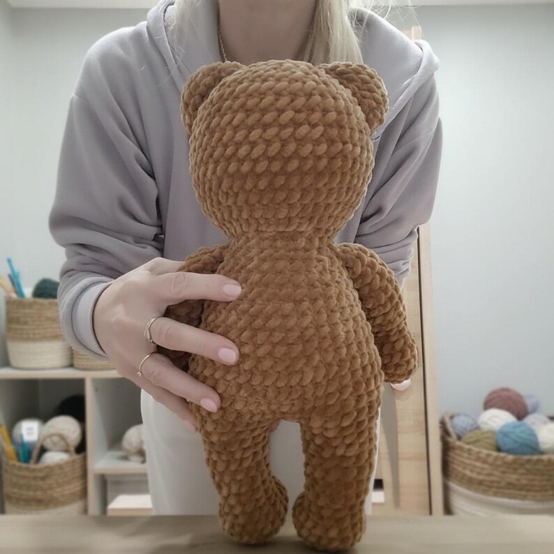 May include: A handmade brown teddy bear made of soft, textured yarn. The bear has a round head, small ears, and a plump body. It is held up by a person. The bear is a classic toy and a cozy gift.
