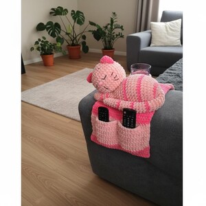 May include: A pink and white striped crochet sofa armrest organizer with two pockets, holding two black remote controls. A plush, sleeping cat-shaped pillow is attached. The organizer is on a gray sofa.