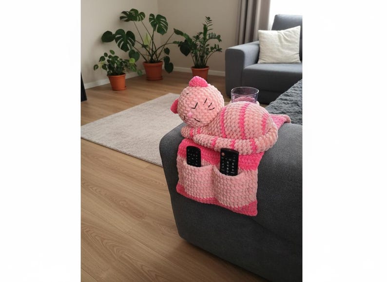 May include: A pink and white striped crochet sofa armrest organizer with two pockets, holding two black remote controls. A plush, sleeping cat-shaped pillow is attached. The organizer is on a gray sofa.
