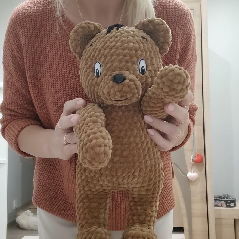 May include: A brown crocheted teddy bear with a friendly expression. The bear has black eyes, a black nose, and a small black patch of hair. The bear is made of a soft, textured yarn and has its right arm raised.