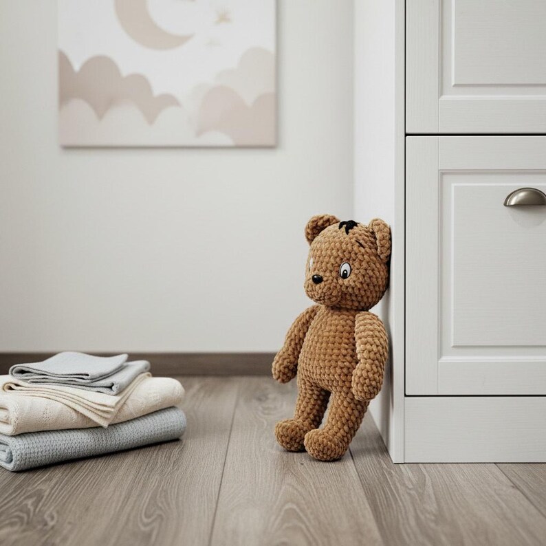 May include: A brown crocheted teddy bear leans against a white cabinet. The bear has black eyes and a small black patch on its head. A stack of folded blankets in shades of gray and cream rests on the wooden floor.