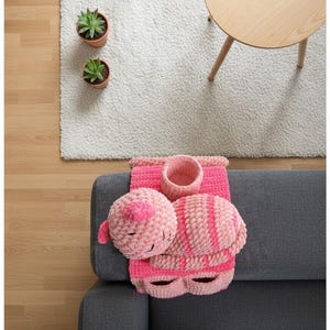 May include: A handmade pink crochet cat-themed set, including a round head, a striped body, and a container. The set is placed on a gray sofa, with a white rug, two potted plants, and a small wooden table in the background.