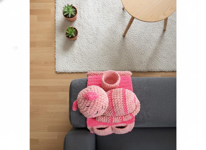 May include: A handmade pink crochet cat-themed set, including a round head, a striped body, and a container. The set is placed on a gray sofa, with a white rug, two potted plants, and a small wooden table in the background.