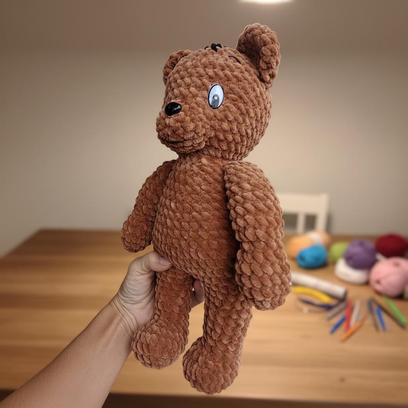 May include: A brown teddy bear made of soft, textured material. The bear has a black nose, a stitched mouth, and a single eye with a blue iris. The bear is approximately 12 inches tall.