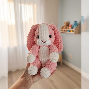 Puede incluir: Un conejito de peluche rosa y blanco hecho a mano con orejas largas, ojos negros y una raya blanca en la cabeza. El conejito está hecho de hilo suave y texturizado y tiene patas y vientre blancos. Un peluche lindo y tierno.