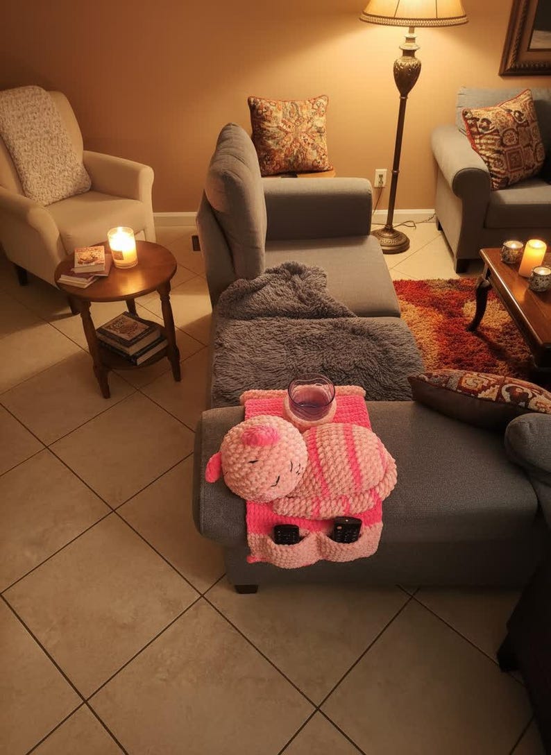 May include: A cozy living room scene featuring a gray sectional sofa with a pink and white crocheted organizer. The organizer holds a glass of purple liquid and two remote controls. A crocheted pillow with a sleeping face is attached.