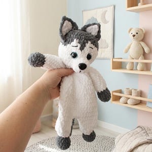 May include: A handmade white and gray husky dog plush toy with black eyes and a black nose. The plush has gray accents on the ears, paws, and tail. The toy is held up in front of a light-colored background.