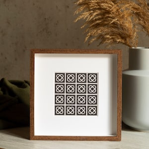 Puede incluir: Un marco de madera marrón con un paspartú blanco y una impresión geométrica en blanco y negro en el interior. La impresión presenta un patrón repetitivo de pequeños círculos con cuatro pétalos. El marco está sobre una superficie de madera marrón claro.