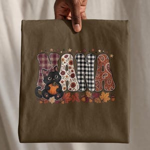 Puede incluir: Una camiseta verde oliva con la palabra NANA en letras grandes estampadas. El diseño incluye un gato negro con un lazo de cuadros sosteniendo una calabaza, rodeado de hojas de otoño, bellotas y pequeñas estrellas. Las letras tienen patrones de cuadros, flores y cachemira.