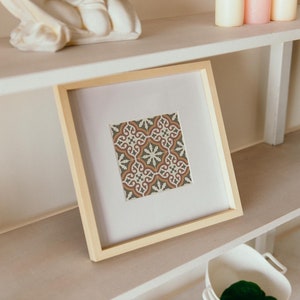 Puede incluir: Una impresión enmarcada de un patrón geométrico de azulejos coloridos en tonos marrones, verdes y blancos. El marco es de madera clara y la impresión está sobre un fondo blanco.