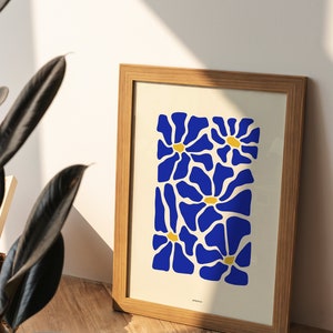 Puede incluir: Un marco de madera rectangular que contiene una impresión sobre un fondo crema. La impresión presenta un patrón repetitivo de flores azules estilizadas con pequeños centros amarillos. La palabra AMEEPIN está impresa en letras negras pequeñas en la parte inferior central.
