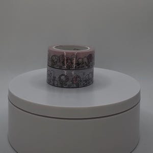 Puede incluir: Dos rollos de cinta adhesiva decorativa apilados. El rollo superior es rosa y el inferior es gris. Ambos rollos presentan un patrón repetido de flores y calaveras de dibujos animados en blanco y negro.