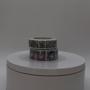 Puede incluir: Dos rollos de cinta adhesiva decorativa apilados sobre una plataforma blanca. El rollo superior presenta un diseño de paisaje urbano en blanco y negro. El rollo inferior muestra personajes de dibujos animados en varios colores.