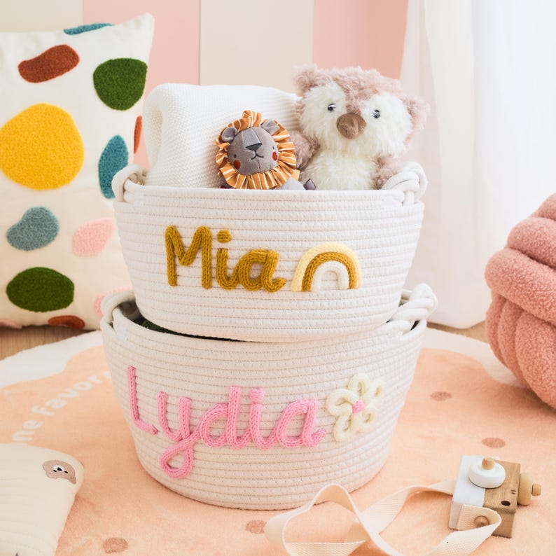 May include: Two white rope baskets stacked on top of each other. The top basket has the name "Mia" in yellow yarn and a rainbow. The bottom basket has the name "Lydia" in pink yarn and a flower. The baskets are filled with toys.