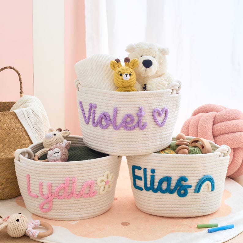 May include: Three cream-colored rope baskets with embroidered names: "Violet", "Lydia", and "Elias". Each basket contains toys, including a teddy bear, giraffe, and wooden toys. The baskets are on a pink rug.