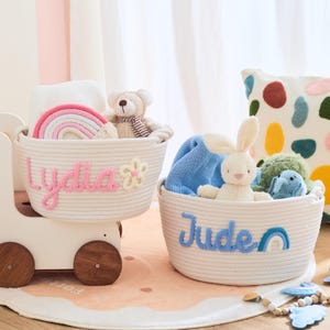 May include: Two white rope baskets with personalized names, "Lydia" in pink and "Jude" in blue, filled with toys. One basket has a rainbow and teddy bear, the other a bunny and elephant. A decorative pillow with colorful spots is in the background.