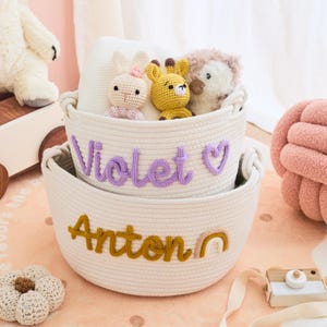 May include: Two off-white rope baskets, one stacked inside the other, with the name "Violet" in purple and "Anton" in gold. The top basket holds stuffed animal toys, including a bunny, giraffe, and hedgehog.
