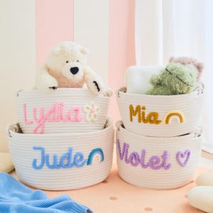 May include: Four white rope baskets with embroidered names and decorative accents. The baskets are stacked, with a teddy bear and stuffed animals inside. Names include "Lydia", "Jude", "Mia", and "Violet".