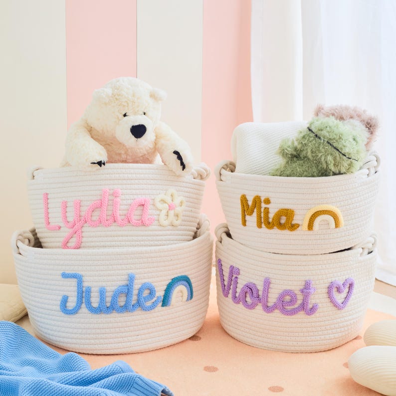 May include: Four white rope baskets with embroidered names and decorative accents. The baskets are stacked, with a teddy bear and stuffed animals inside. Names include "Lydia", "Jude", "Mia", and "Violet".