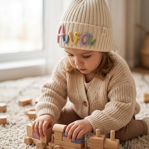 Puede incluir: Un gorro de punto acanalado de color crema con el nombre Ava bordado en letras de hilo en relieve y multicolores en el dobladillo frontal. El gorro tiene un borde doblado y una pequeña etiqueta de tela rectangular en el lateral.