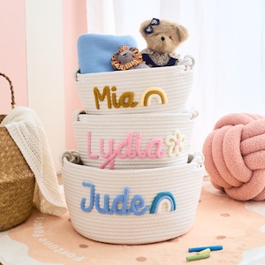 Op de afbeelding: Drie gestapelde, crèmekleurige touwmanden met geborduurde namen: "Mia", "Lydia" en "Jude". Elke mand heeft een decoratief element: een regenboog, een bloem en een regenboog. De bovenste mand bevat een blauwe deken en speelgoed.