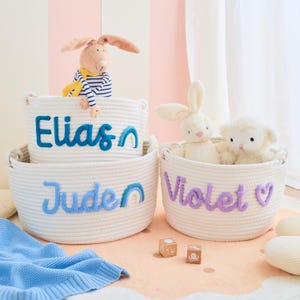 May include: White rope baskets with embroidered names: Elias, Juden, and Violet. Each basket contains a stuffed animal. A blue blanket and wooden blocks are also present. The baskets are on a pink and beige surface.