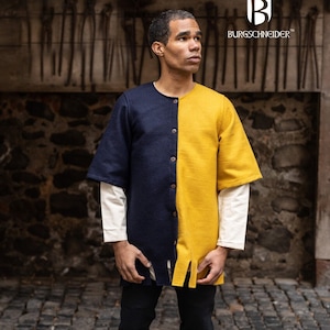 Op de afbeelding: Een persoon draagt ​​een blauwe en gele tuniek met een knoopsluiting. De tuniek heeft een gespleten zoom en is gemaakt van een dikke stof. De persoon draagt ​​een zwarte broek en bruine laarzen.