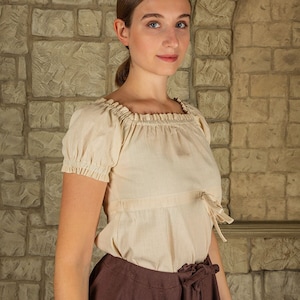 May include: A woman wearing a cream-colored, off-the-shoulder peasant top with short sleeves and a brown skirt. The top has a gathered neckline and ties at the waist.