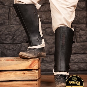 May include: Black leather leg armor with buckles and straps. The armor is worn over white pants and black shoes. The armor is shown on a wooden crate.