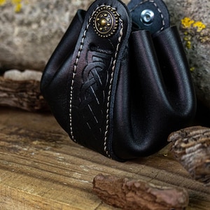 Può includere: Un porta monete in pelle nera con un design a nodo celtico e una chiusura a bottone in ottone. Il porta monete è appoggiato su una superficie di legno.