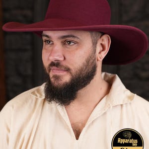 Peut inclure: Un chapeau à large bord en feutre bordeaux avec une couronne arrondie. Le chapeau est porté par une personne avec une barbe. Le chapeau est un style classique avec un large bord qui peut être porté relevé ou abaissé.