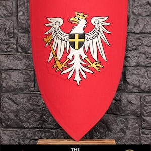 Op de afbeelding: Rood schild met een witte adelaar en een geel kruis op zijn borst. De adelaar draagt een gele kroon op zijn kop. Het schild is uit het videospel "The Witcher".