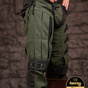 May include: Green and black trousers with a decorative design. The trousers have a black band around the knee with cutouts. The trousers are made of a thick fabric and have a drawstring at the waist.