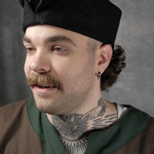 Può includere: Una persona indossa un cappello di feltro nero e marrone con una falda appuntita sul davanti. Il cappello è indossato basso sulla fronte. La persona ha i baffi e un tatuaggio sul collo.