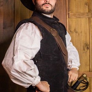 Peut inclure: Un homme portant un chapeau noir à larges bords, une chemise blanche, un gilet en cuir noir avec une sangle en cuir marron et une boucle en laiton, et un pantalon noir. Il tient une épée avec une poignée dorée. Le texte "RAWBLADE EAST COAST MERCHANTS EST. 2018" est visible dans le coin inférieur droit de l'image.